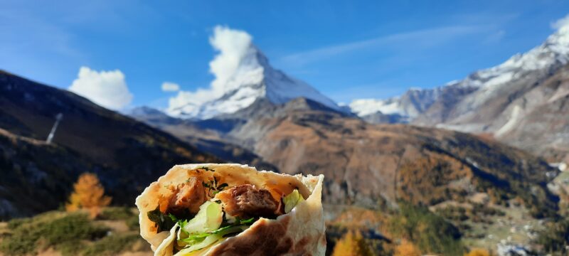 Na imagem, em primeiro plano um burrito vegano recheado com guacamole, ao fundo Matterhorn na Suíça.